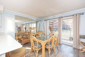 Ein Essbereich mit einem Glastisch, umgeben von Korbstühlen, einer weißen Theke und Blick auf einen Balkon mit Vorhängen.