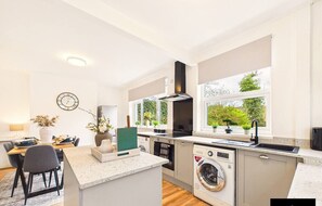 Een moderne keuken met een ingebouwde wasmachine, een eethoek met een tafel en stoelen, en grote ramen met uitzicht op groen.