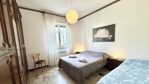 Un dormitorio con cama, una silla, una mesita de noche, una lámpara, una ventana con cortinas y un cuadro colgado en la pared.