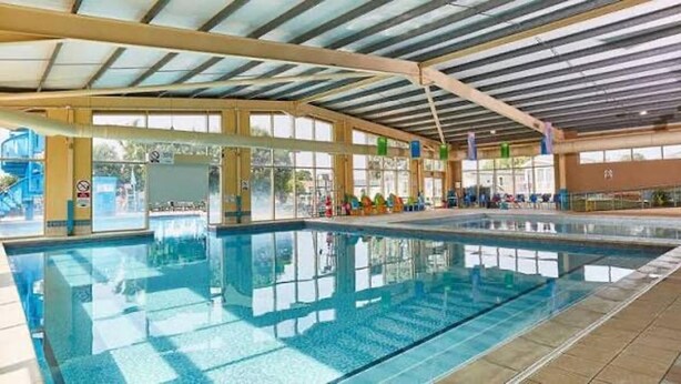 Una amplia piscina interior con agua cristalina, rodeada de ventanales y un techo alto con vigas metálicas.
