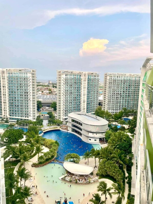 A high-rise hotel with multiple balconies, a central swimming pool, and a beach area with palm trees.