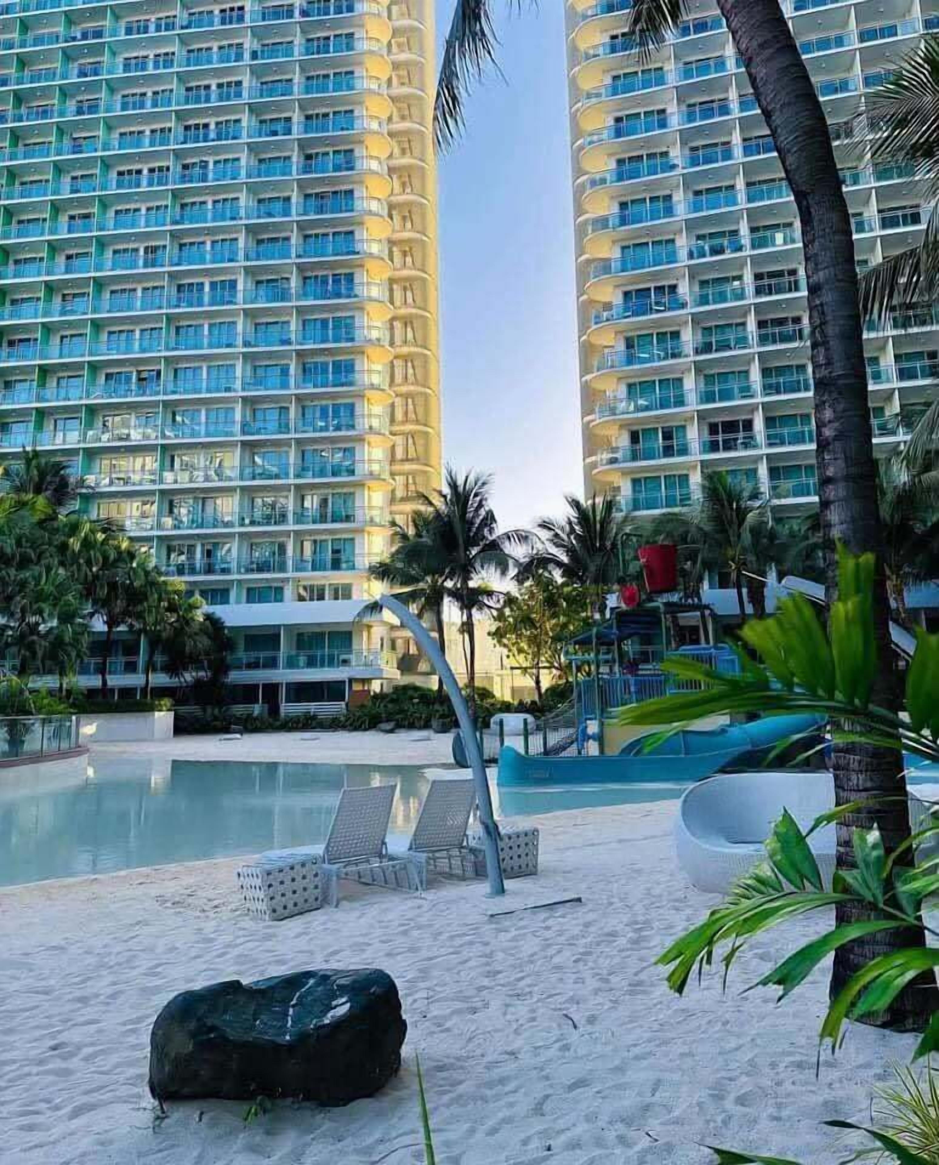 Een resort met een zwembad, ligbedden, palmbomen en hoge gebouwen op de achtergrond.