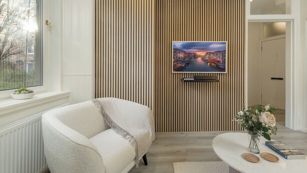 Una sala de estar moderna con un sofá, un televisor de pantalla plana montado en un panel de madera, un jarrón con flores y una ventana con vistas a edificios.