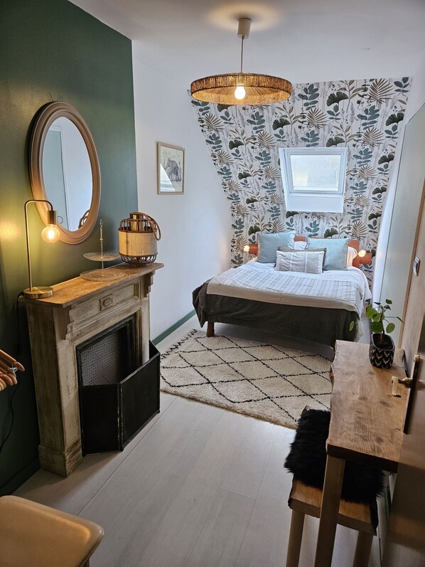 A bedroom with a bed, a bedside table, a mirror, a hanging light fixture, a fireplace, and a window.