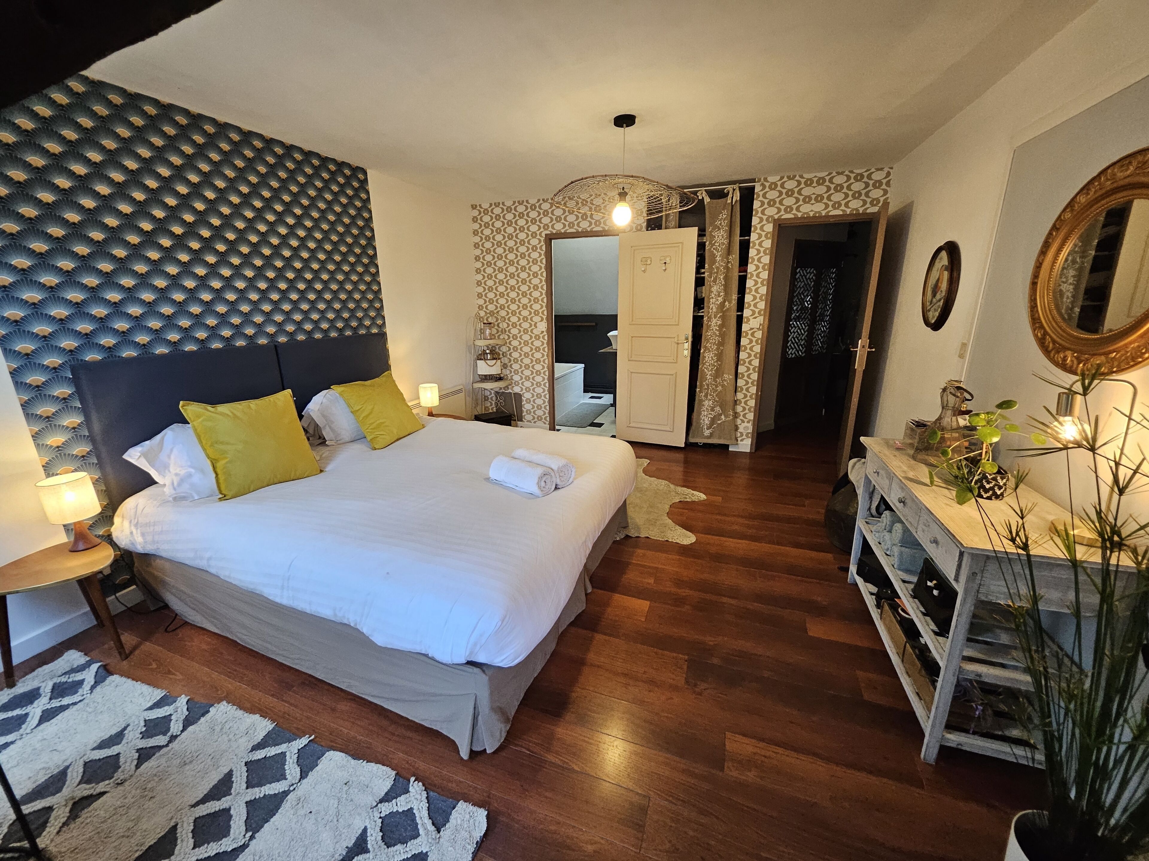 A bedroom with a bed, bedside lamps, a wooden dresser, a mirror, and a patterned wall.