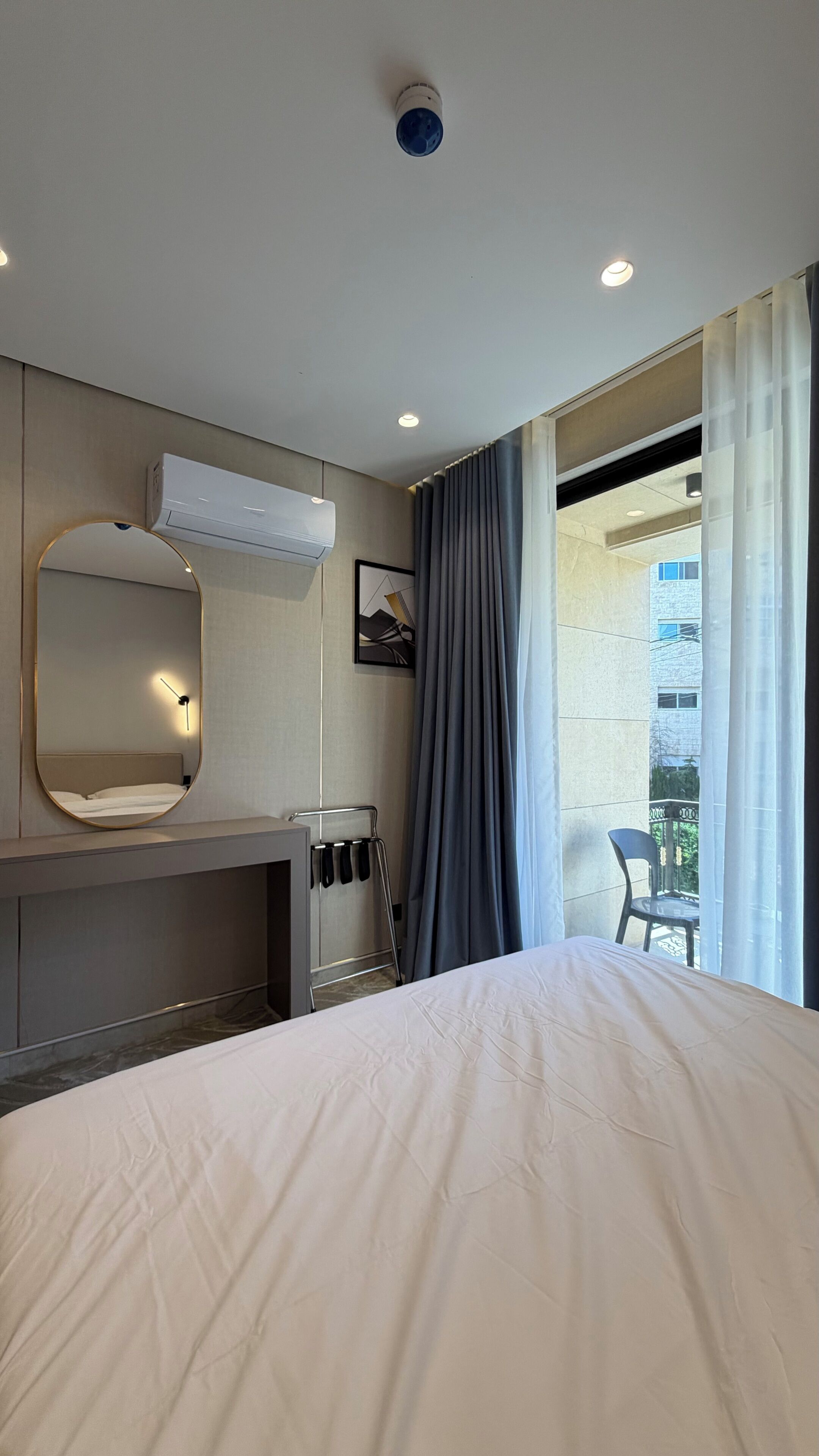 Una habitación de hotel con cama, espejo, radiador, ventana con cortinas y equipo de aire acondicionado.