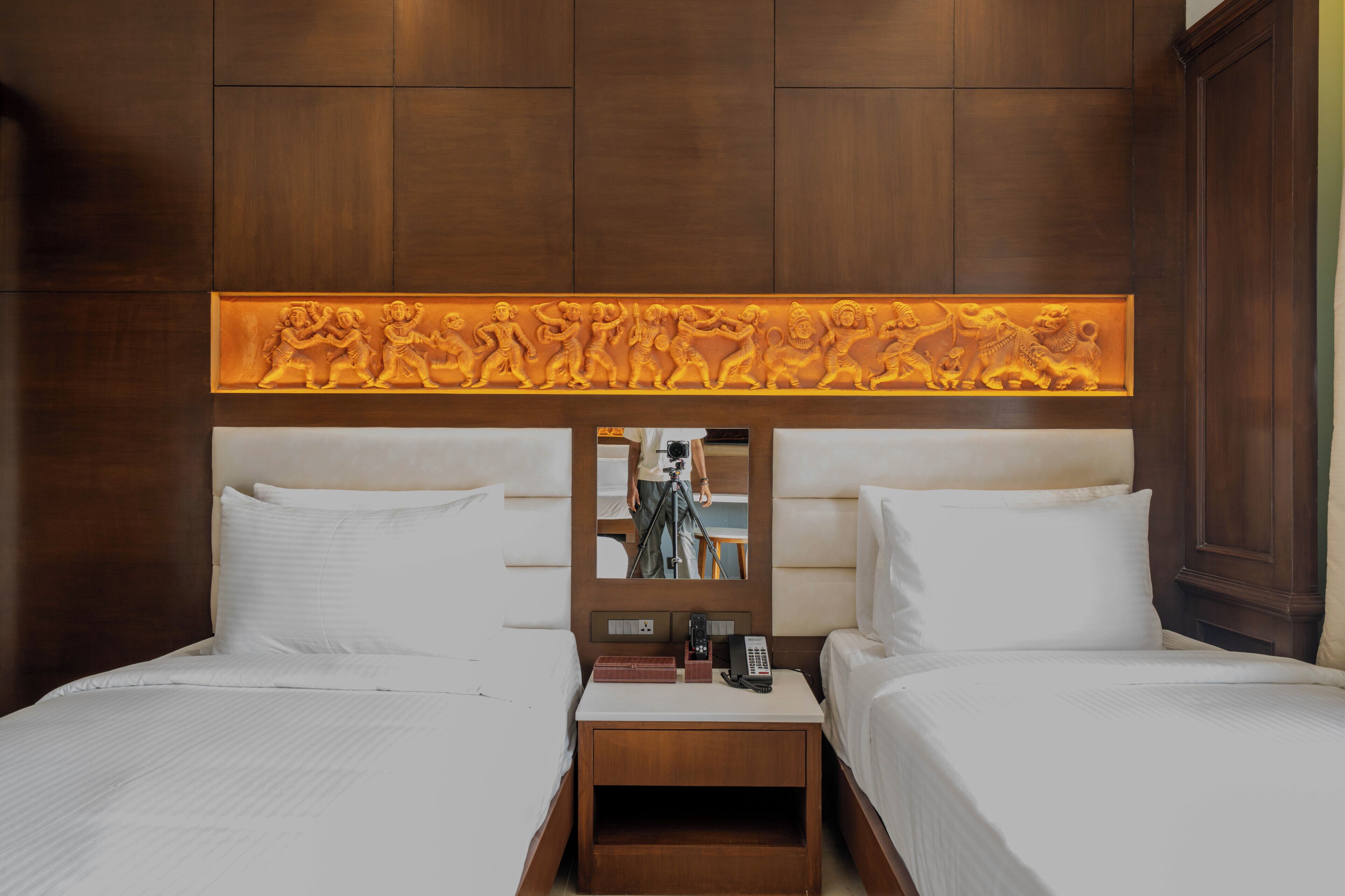 Habitación de hotel con dos camas, cabecero de madera con un relieve decorativo, mesita de noche con lámpara y espejo.