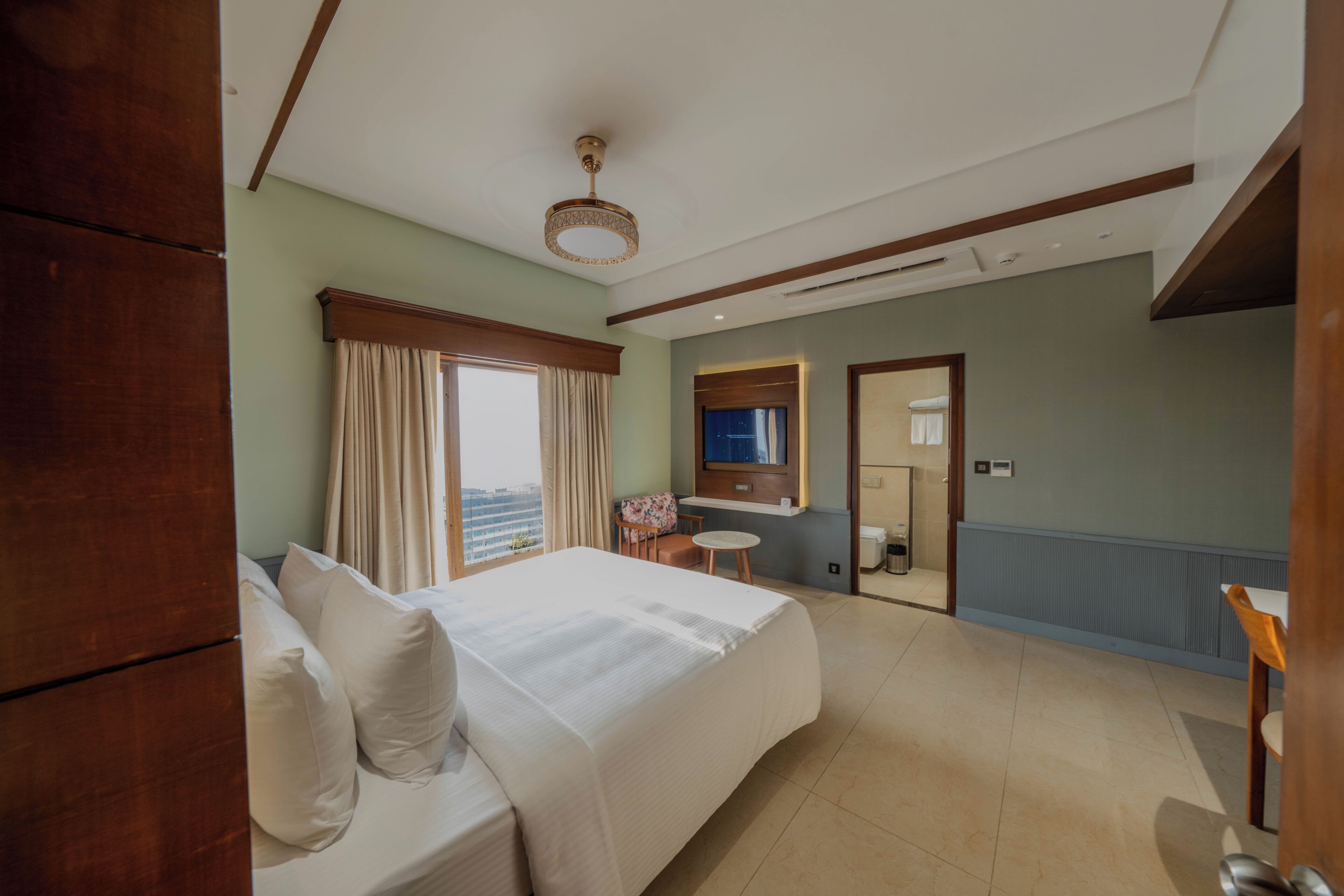 A hotel room with a large bed, a small table, a chair, a mirror, and a view of the city.