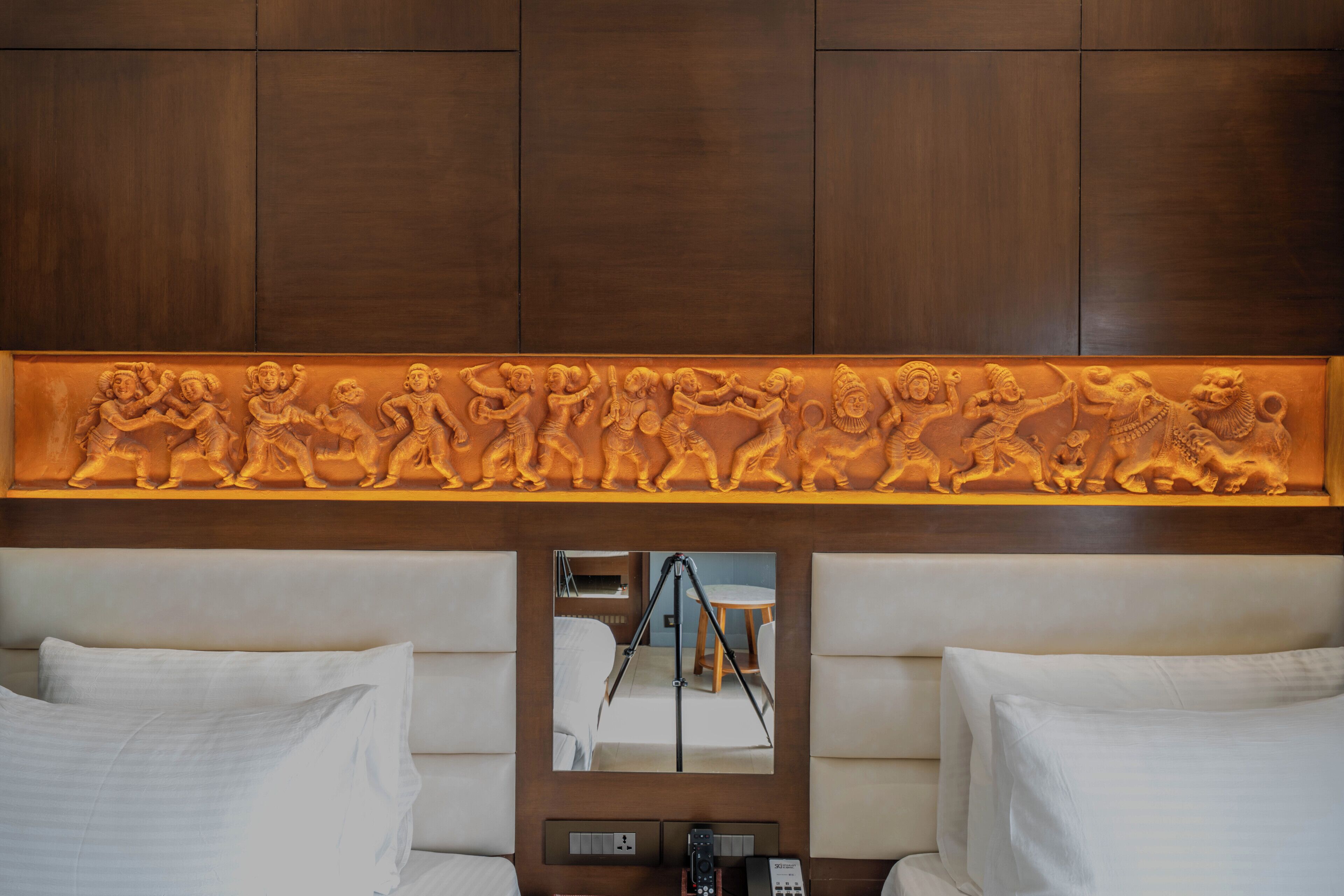 A hotel room with a carved wooden panel above the bed, a mirror, and a control panel with a phone.
