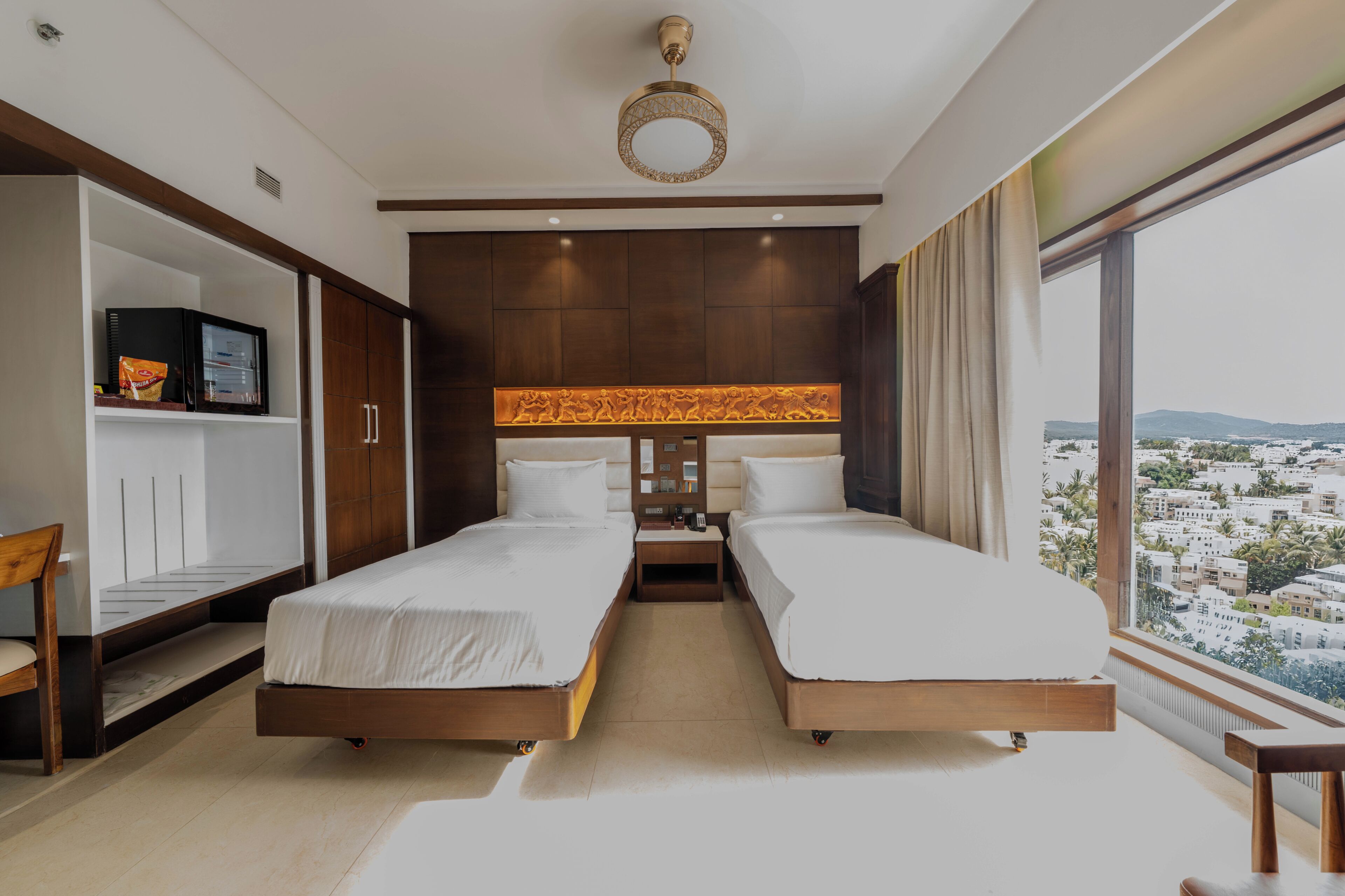 A hotel room with two beds, a TV, a minibar, and a large window with a city view.