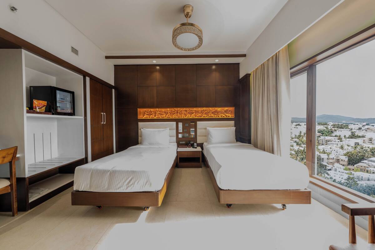 A hotel room with two beds, a TV, a minibar, and a large window with a city view.