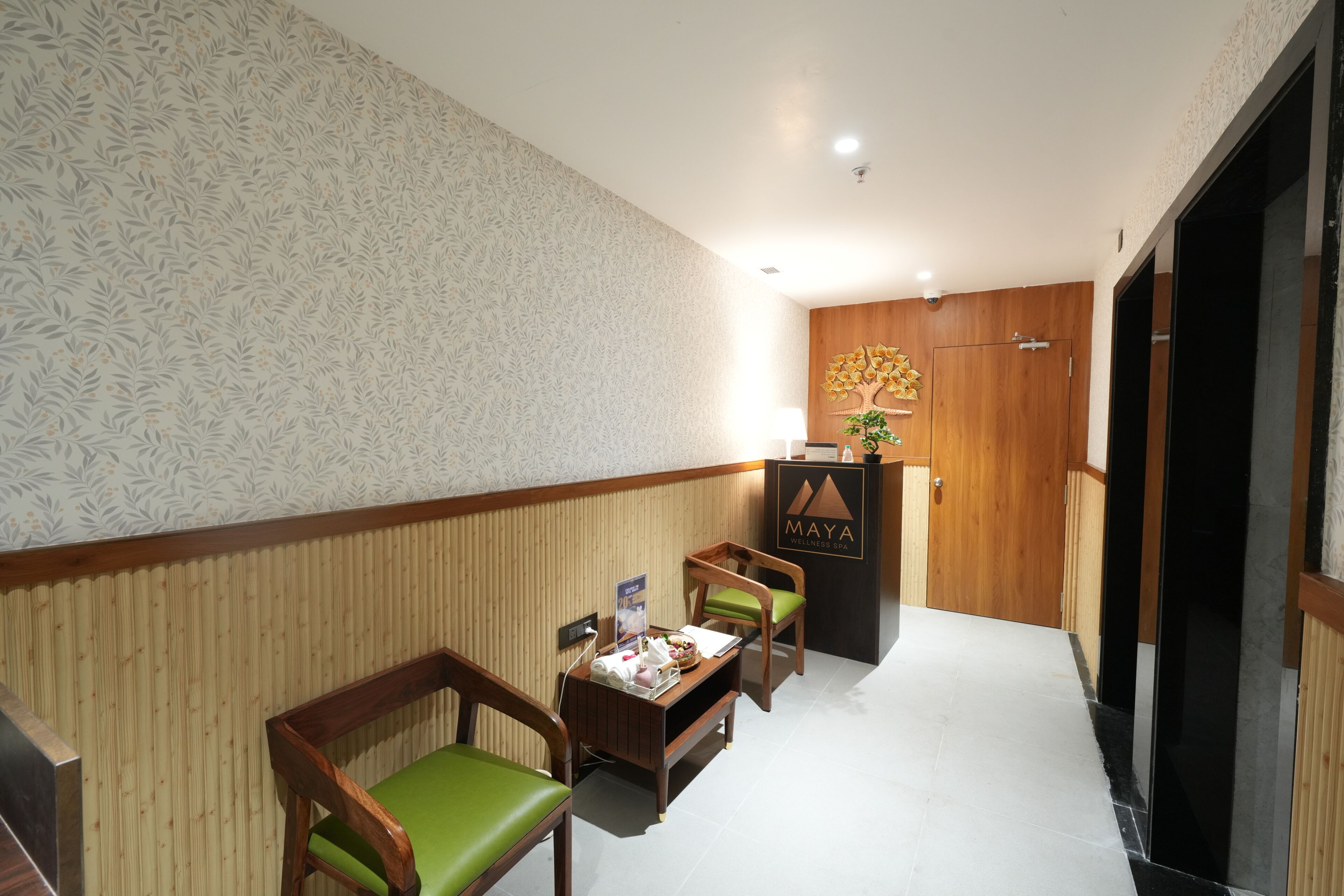 A hotel lobby with a reception desk, wooden paneling, a green cushioned chair, a small table with a tray, and a wall-mounted plant.