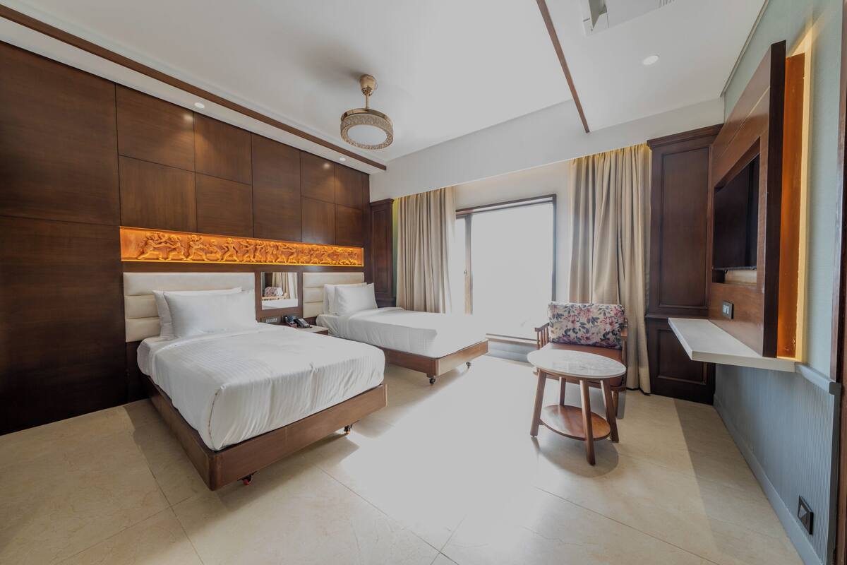 Habitación de hotel con dos camas, una mesita, una silla, televisor y ventana con cortinas.