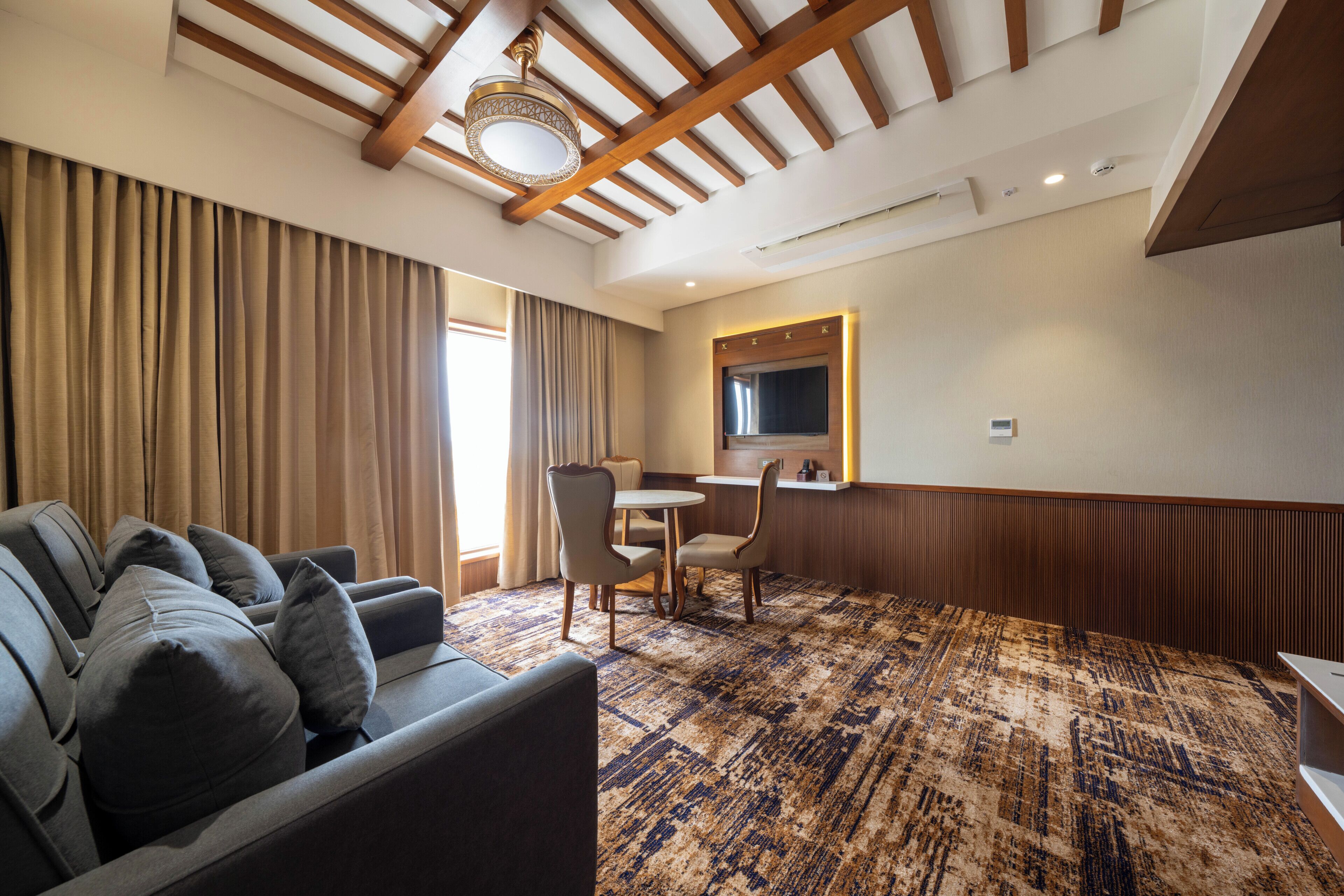 A hotel room with a sofa, a dining table with chairs, a TV mounted on the wall, and a patterned carpet.