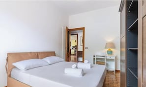 Habitació d'hotel amb un llit, una taula de nit amb una làmpada, un armari i una porta que condueix a una altra habitació.