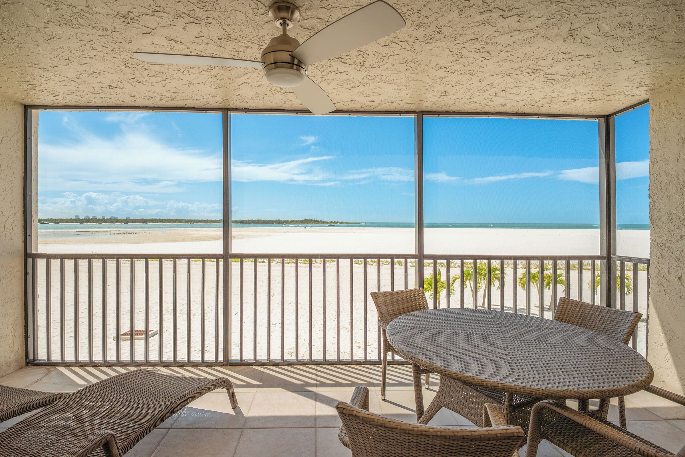 A balcony with a view of a beach, a ceiling fan, and wicker furniture.