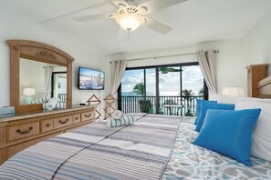 Ein Schlafzimmer mit einem großen Bett, einem Deckenventilator, einem Fernseher, einem Balkon mit Aussicht und einer Holzkommode.