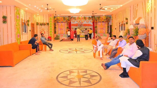 A room with orange sofas arranged in a semi-circle, a chandelier, ceiling fans, and a decorative wall with a star pattern. - Shree Videhi Atithi Grah (Samudrapur)