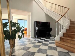 Ein geräumiger Wohnbereich mit Schachbrettmuster-Boden, einer Treppe, einem Klavier und einer großen Pflanze.