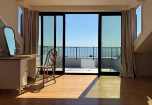 Ein Zimmer mit Holzboden, einem Schreibtisch mit Stuhl und einer großen Schiebetür, die auf einen Balkon mit Meerblick führt.