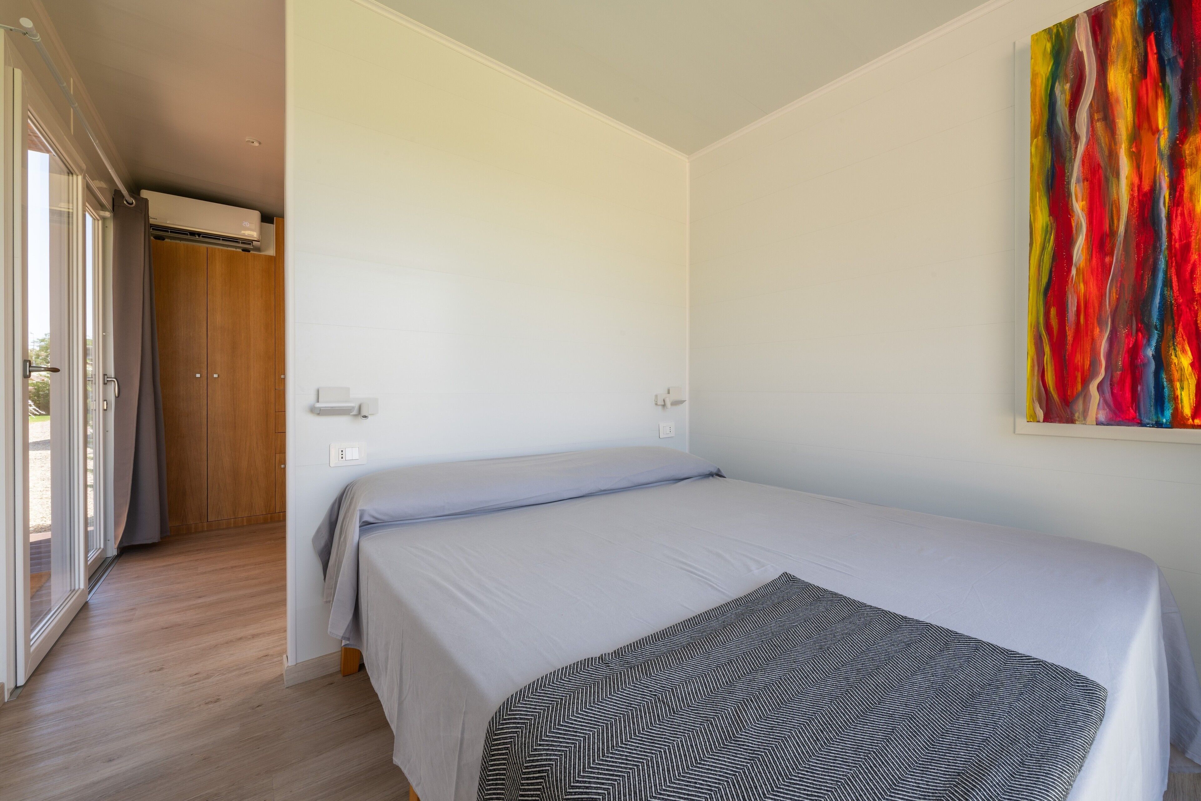 A bedroom with a bed, a door leading to a balcony, and a colorful abstract painting on the wall.