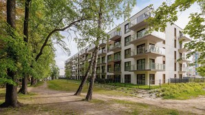 Een meerlaags woongebouw met balkons, omgeven door bomen en een onverhard pad.