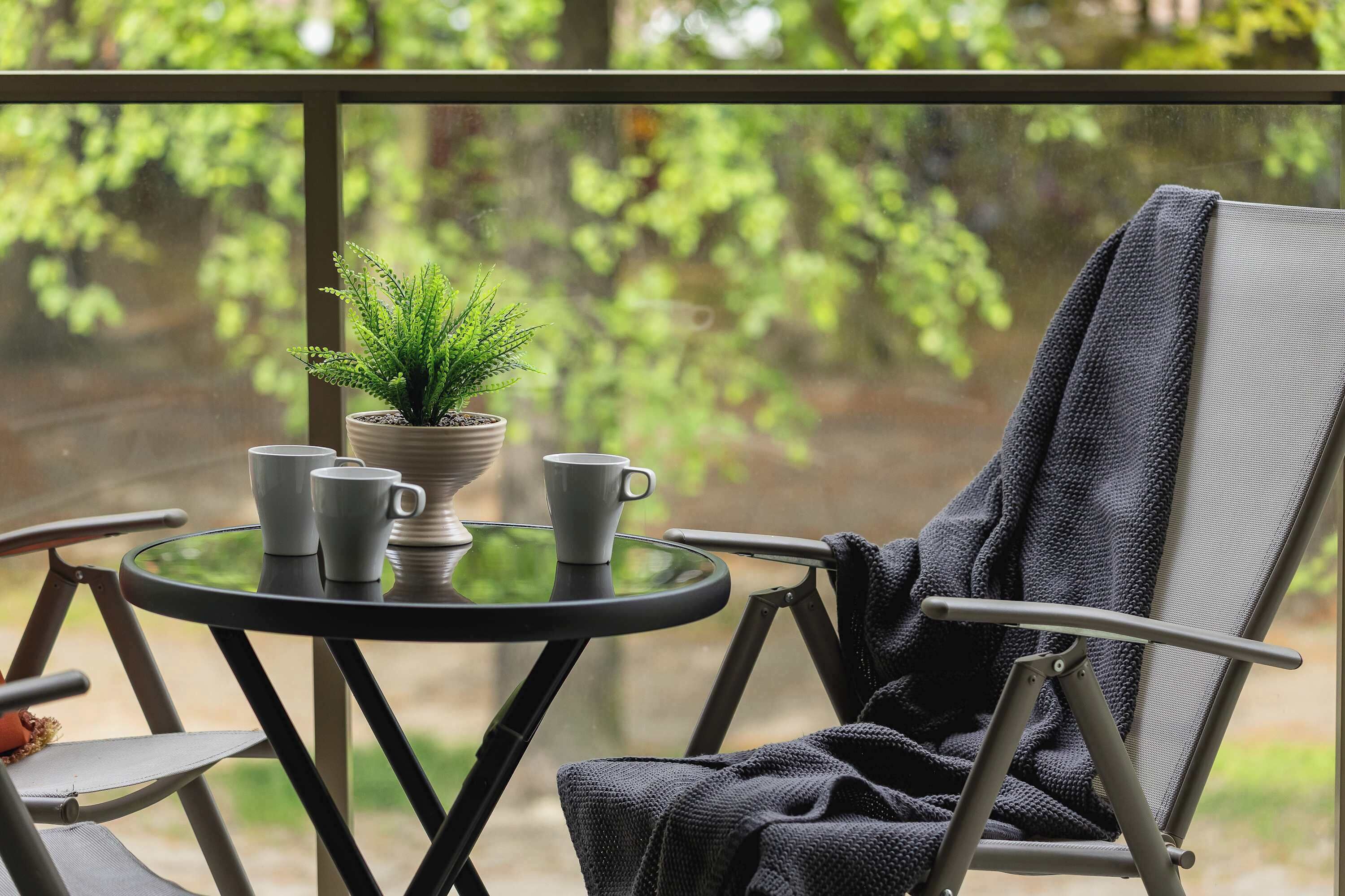 A balcony with a table, chairs, a potted plant, and a window with a view of greenery.