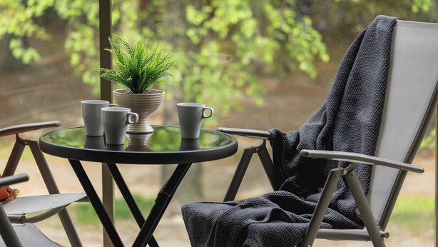 Een balkon met een tafel, stoelen, een bloempot en een raam met uitzicht op groen.