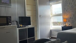 A hotel room with a flat-screen TV on a white stand, a small desk with a kettle and a coffee maker, a bed with a grey headboard, a window with a floral wallpaper, and a nightstand with a lamp and a bottle.