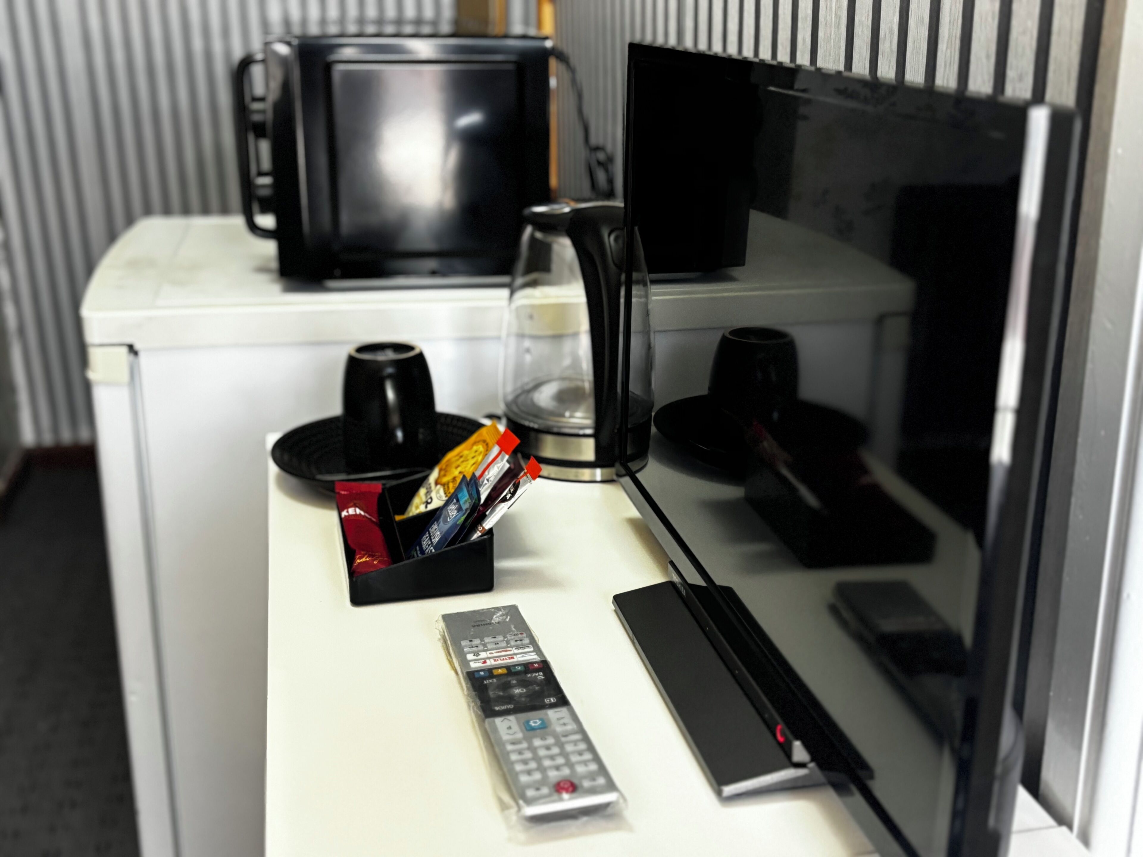 A hotel room with a white desk, a remote control, a glass of water, a coffee maker, and a microwave.