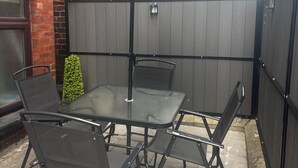 A patio with a glass table and four chairs, a large umbrella, and a brick wall.