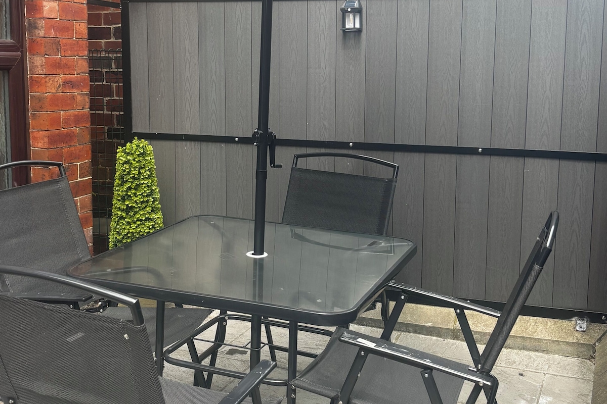 Un patio con una mesa de vidrio, sillas de metal, un paraguas oscuro y una cerca de madera.