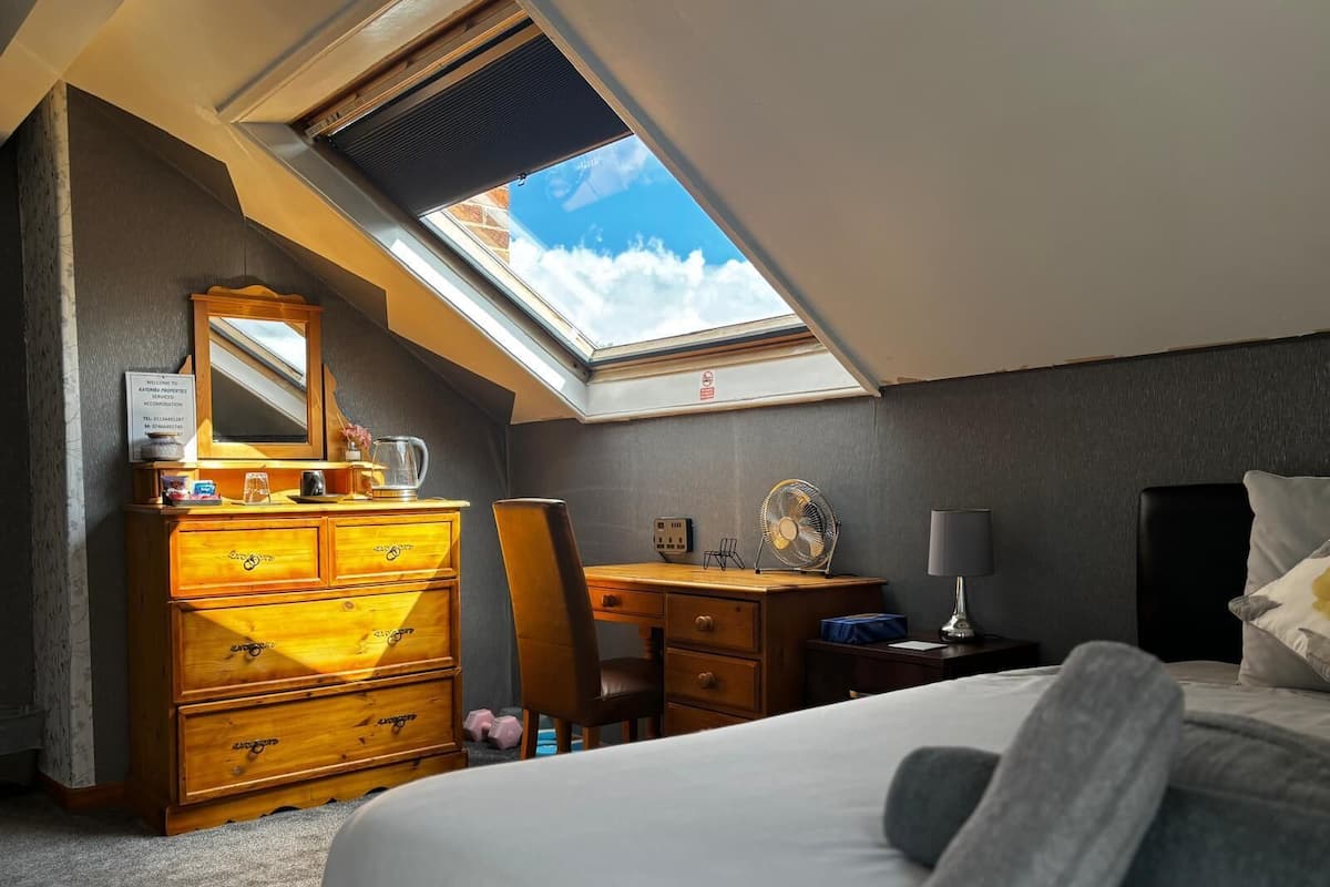 A bedroom with a skylight, a wooden dresser, a desk, a chair, a bed, and a nightstand.