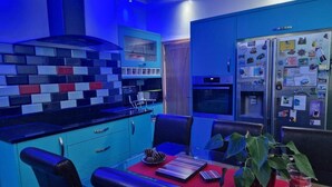 A modern kitchen with blue cabinets, a red dining table, and a refrigerator with magnets.