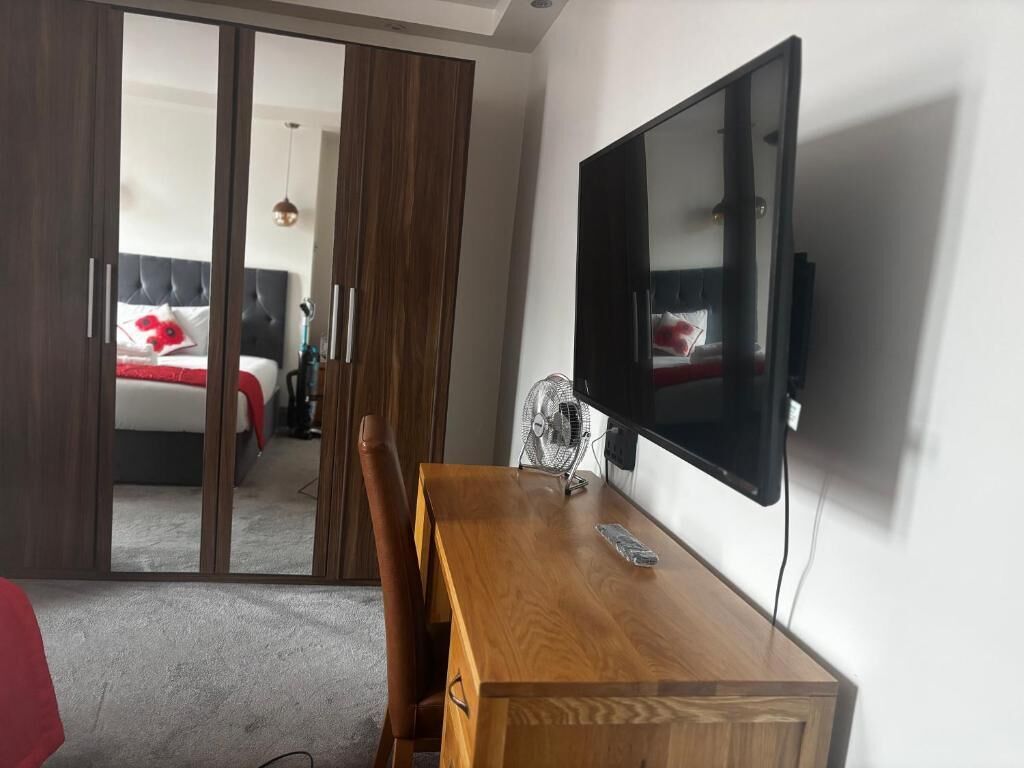 A room with a wooden dining table, a flat-screen TV mounted on the wall, a sliding door leading to a bedroom with a bed and a red pillow.