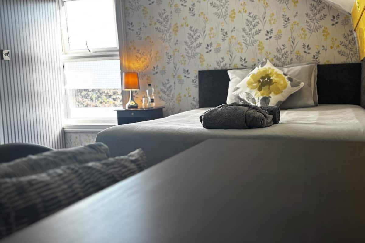A neatly made bed with a pillow and a folded towel, a bedside lamp, and a floral wallpaper pattern.