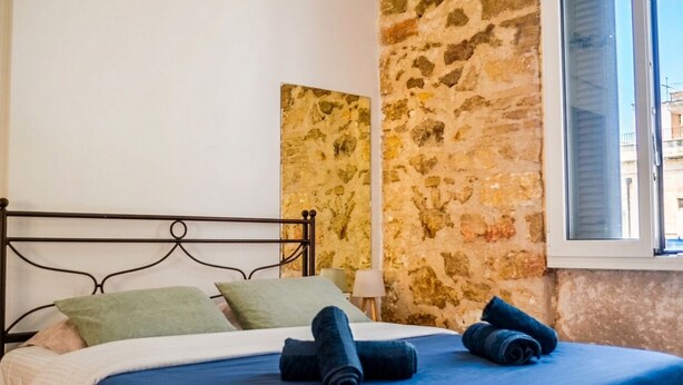 A bed with a metal frame, a blue bedspread, and rolled towels. There is a stone wall and a window with blinds. - Kerameikos Radio-Apartments (Athina)
