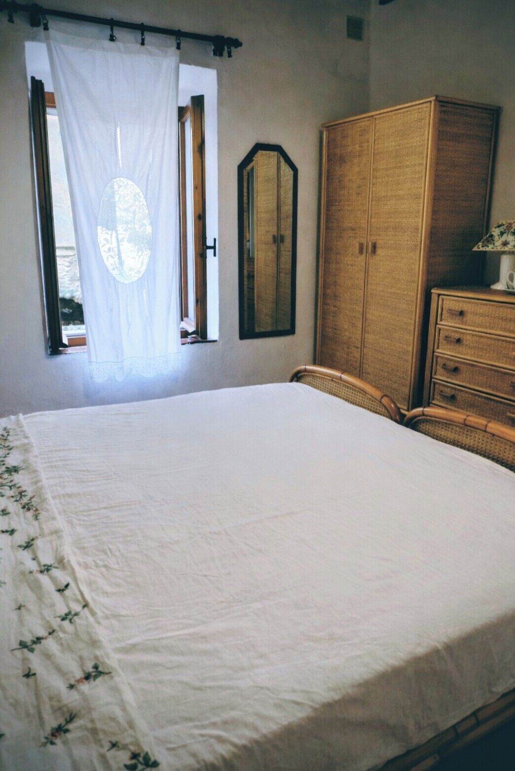 A bedroom with a large bed, a wooden wardrobe, a mirror, and a window with curtains.