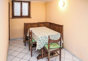 Un comedor con una mesa dispuesta para cuatro, sillas de madera con cojines verdes, mantel con estampado floral, una ventana con cortinas de encaje y una lámpara fijada a la pared.