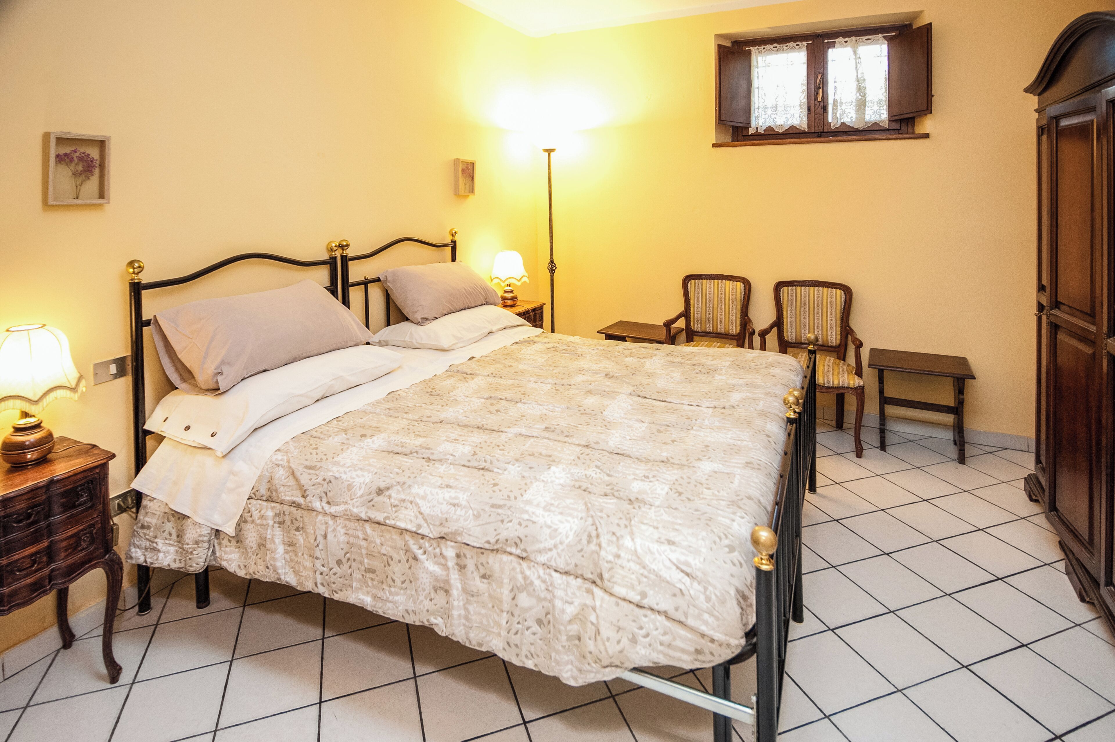 A bed with a patterned bedspread, a bedside lamp, a nightstand, two chairs, a small table, and a wardrobe in a room with tiled flooring and a window with curtains.