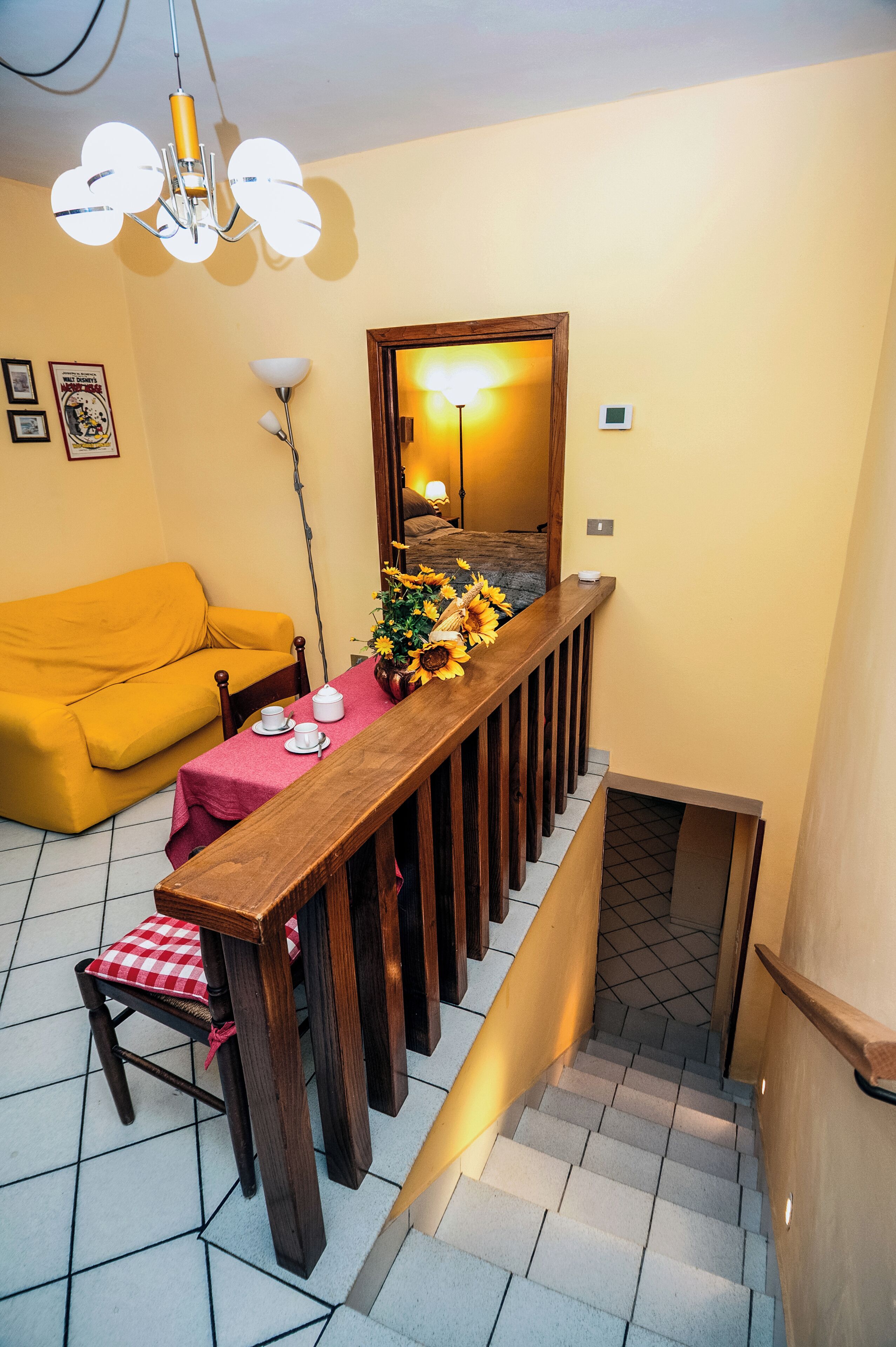 A staircase with a wooden railing, a yellow sofa, a dining table with a red tablecloth, and a vase with sunflowers.