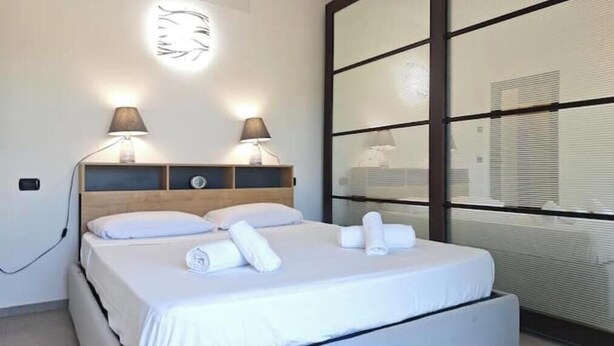 A hotel room with a bed, two bedside lamps, a wall-mounted light, and a large window with blinds. - Hostdomus - Azimut Apartment (Liguria)