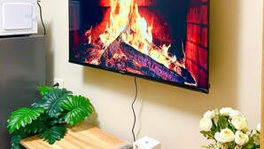 A flat-screen TV mounted on a wall, a wooden table with a remote, a white box, and a shelf with plants. - Cozy 1-bedroom condo walking distance to MOA (Pasay City)