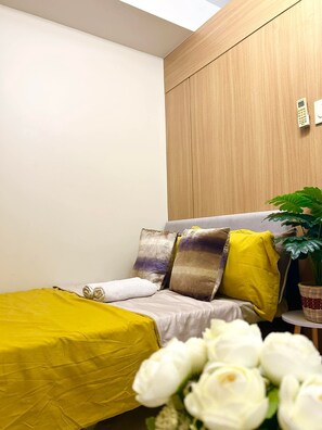 A bedroom with a bed, pillows, a bedside table, a plant, and a wall-mounted light switch. - Cozy 1-bedroom condo walking distance to MOA (Pasay City)