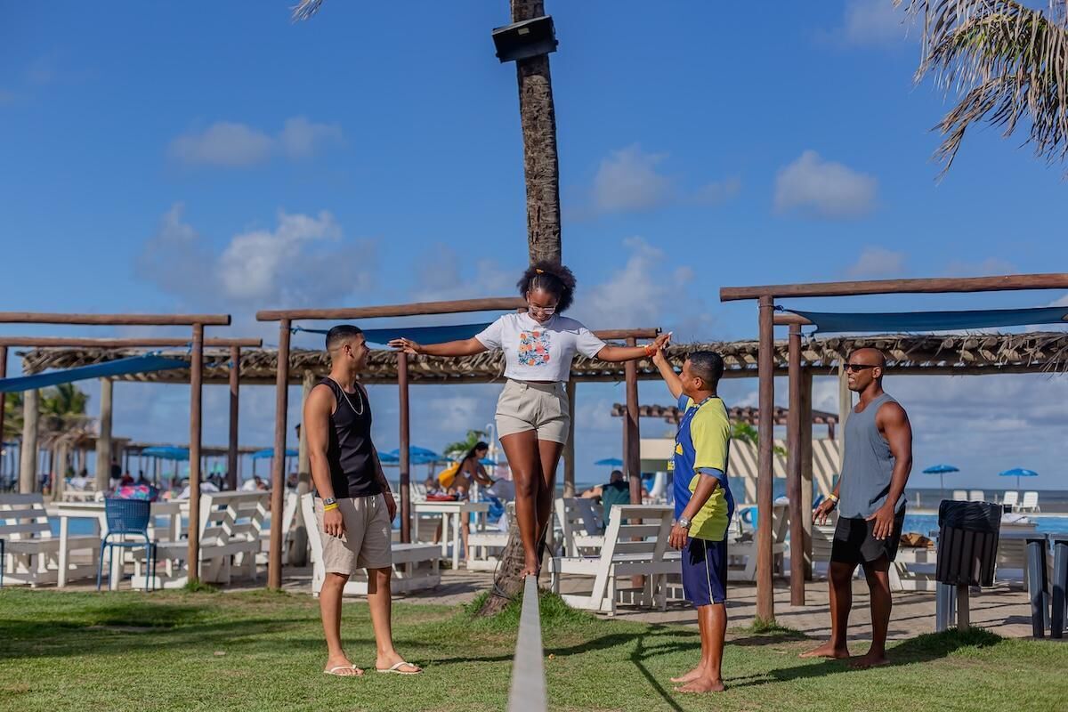 Cenário à beira-mar com uma pessoa equilibrando-se num mastro, cercada por cadeiras de praia e guarda-sóis.