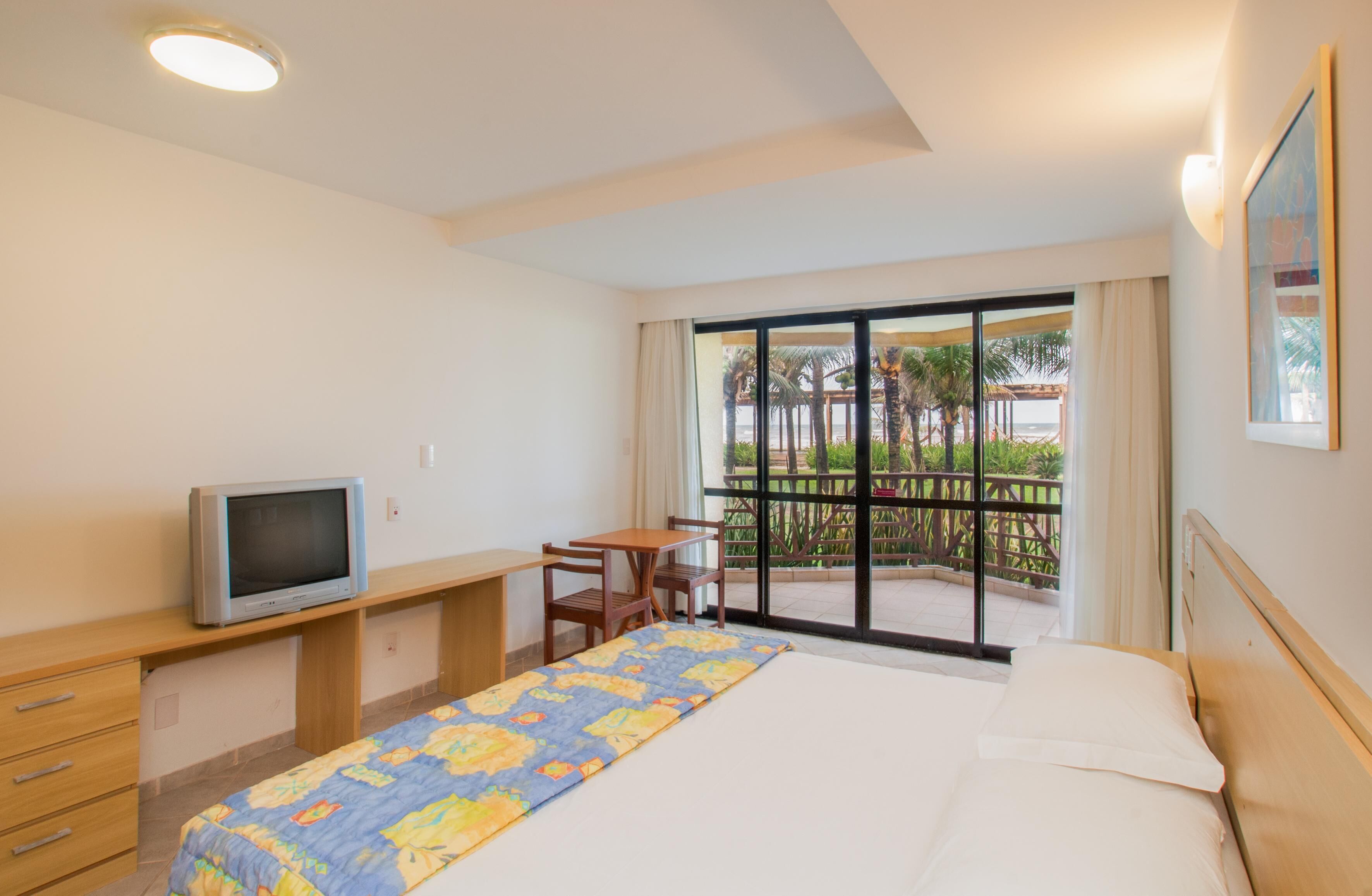 A hotel room with a bed, a desk, a television, and a balcony with a view of palm trees and a beach.