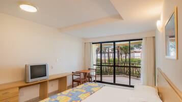 A hotel room with a bed, a desk, a television, and a balcony with a view of palm trees and a beach.