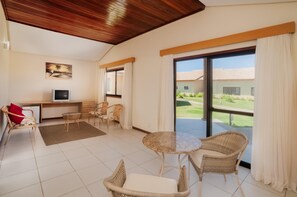 A spacious room with a large sliding glass door leading to a balcony, a small desk with a television, wicker furniture, and a painting on the wall.