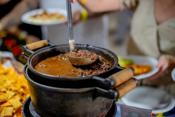 A large pot of stew with a ladle, surrounded by plates of food and a bowl of chips.