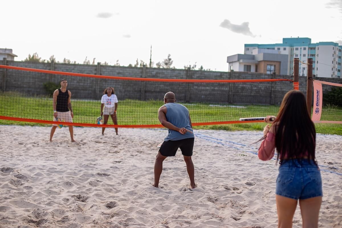 A beach volleyball game with players on a sandy court, a net, and a building in the background.
